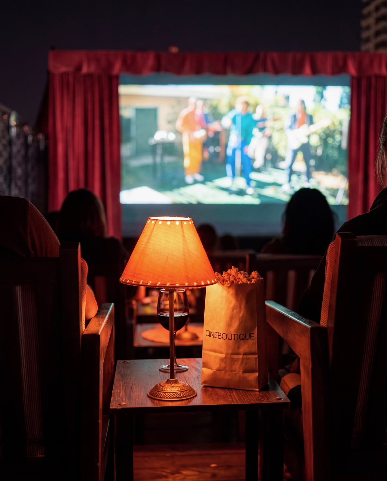 Una lámpara encendida, una copa de vino y pochoclo de Cine Boutique sobre una mesa, frente a la pantalla entre cortinas rojas en la terraza